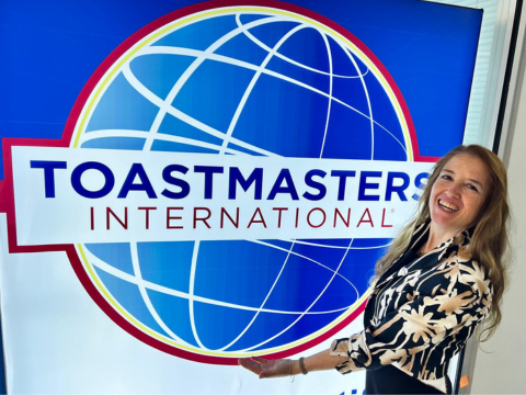 Deniz is smiling at camera and holds a Toasmasters flag