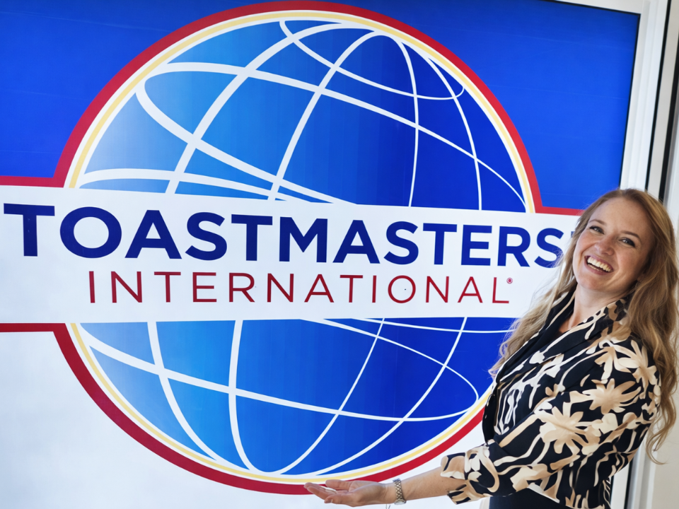 Deniz is smiling at camera and holds a Toasmasters flag